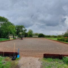 A wide view of a horse riding arena surrounded by trees under a cloudy sky. A rider on horseback is visible in the distance. The foreground shows a path leading to the arena with gardening tools nearby.