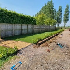 A sunny outdoor scene with freshly laid gravel and wooden borders separating a grassy area from a gravel path. A white van is parked nearby, and a person stands next to it. Tall trees and a wooden fence line the background.