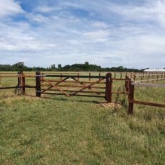 A wooden farm gate stands open, leading to a vast, grassy field. Fence lines extend into the distance under a partly cloudy sky. Trees and a few buildings are visible on the horizon.