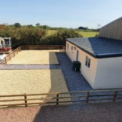 A small, white prefabricated building with a black roof is situated next to a wooden-fenced area. There are two gravel sections beside it, and a digger is in the background with some workers nearby. Trees and open fields surround the scene.