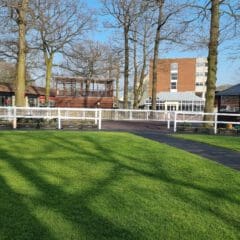 A sunny day at an outdoor venue featuring a circular path surrounded by green grass and tall leafless trees. Buildings with large windows and a red brick structure are in the background, under a clear blue sky.