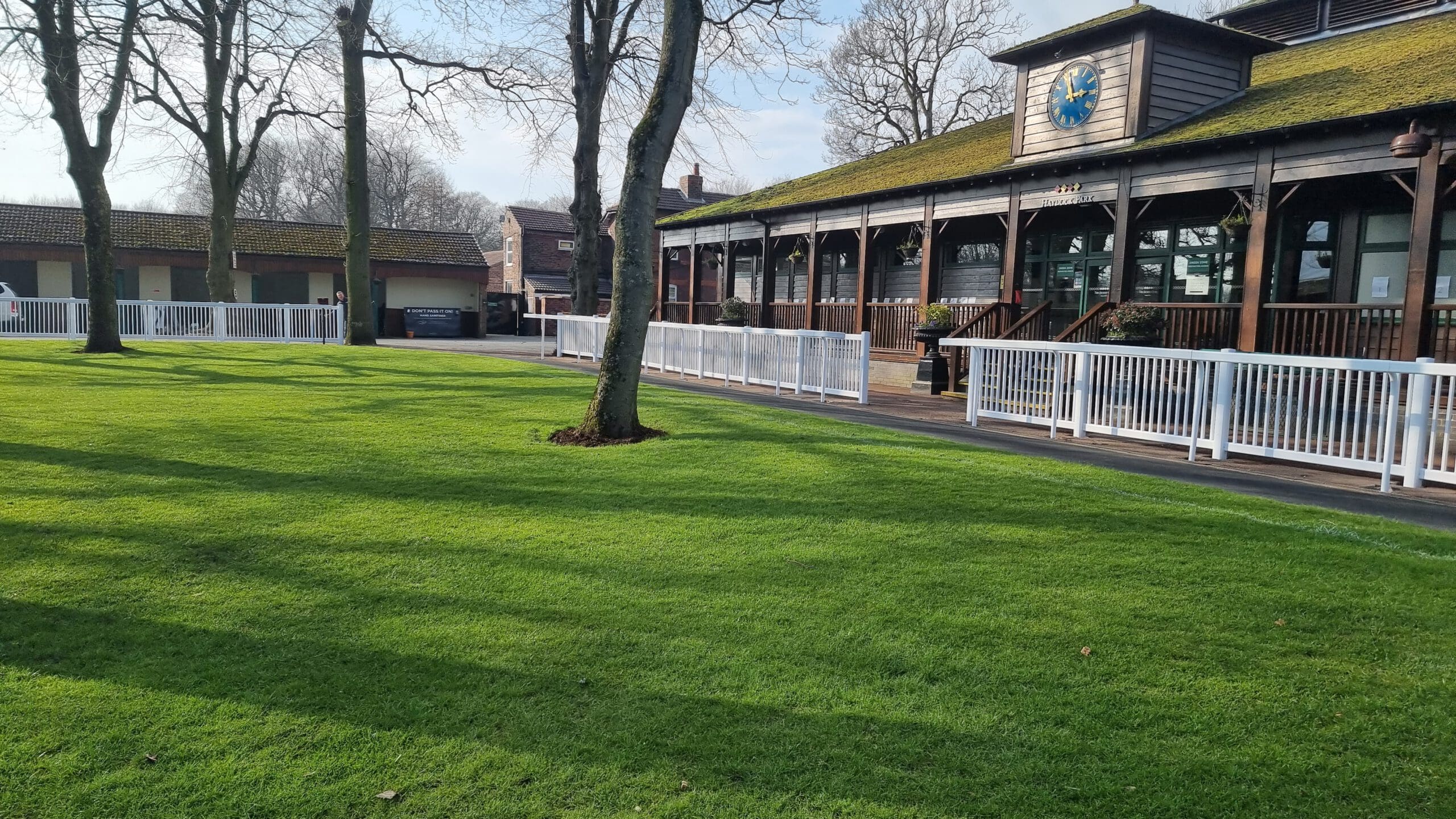 A well-maintained grassy area with a few trees, adjacent to a wooden building featuring a turquoise clock. A white fence lines the pathway beside the building, and there are more structures visible in the background.