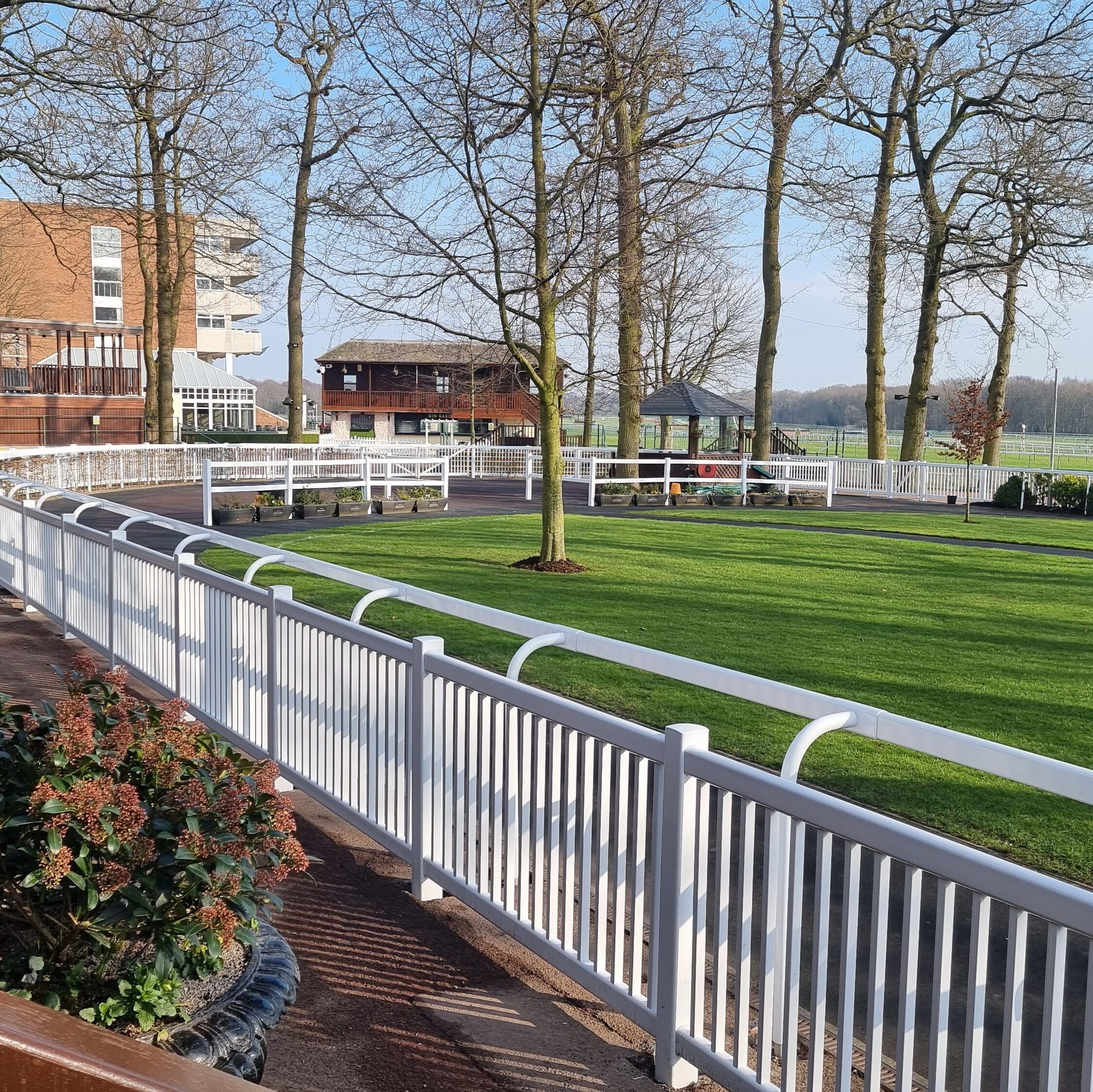 View of a serene, fenced horse paddock with neatly trimmed grass, surrounded by tall trees. Buildings are visible in the background and a few red and green potted plants are in the foreground. The sky is clear and blue.