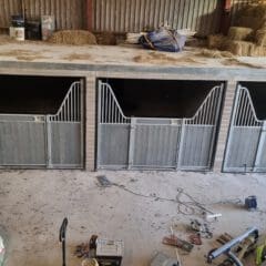 A stable with three empty horse stalls, featuring metal gates. The floor is scattered with tools, bags, and equipment, and hay bales are stacked above. The setting appears to be a work in progress.