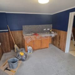 A room with dark blue and wooden paneled walls, featuring a tiled floor. A wooden cabinet with a plastic cover underneath a grey tiled backsplash is against the wall. A ladder is visible in the doorway to another room. Tools and buckets are on the floor.