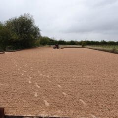 A large, rectangular outdoor equestrian arena with sandy ground and hoofprints. A tractor is seen in the distance preparing the surface. The area is surrounded by lush green trees and open fields under a cloudy sky.