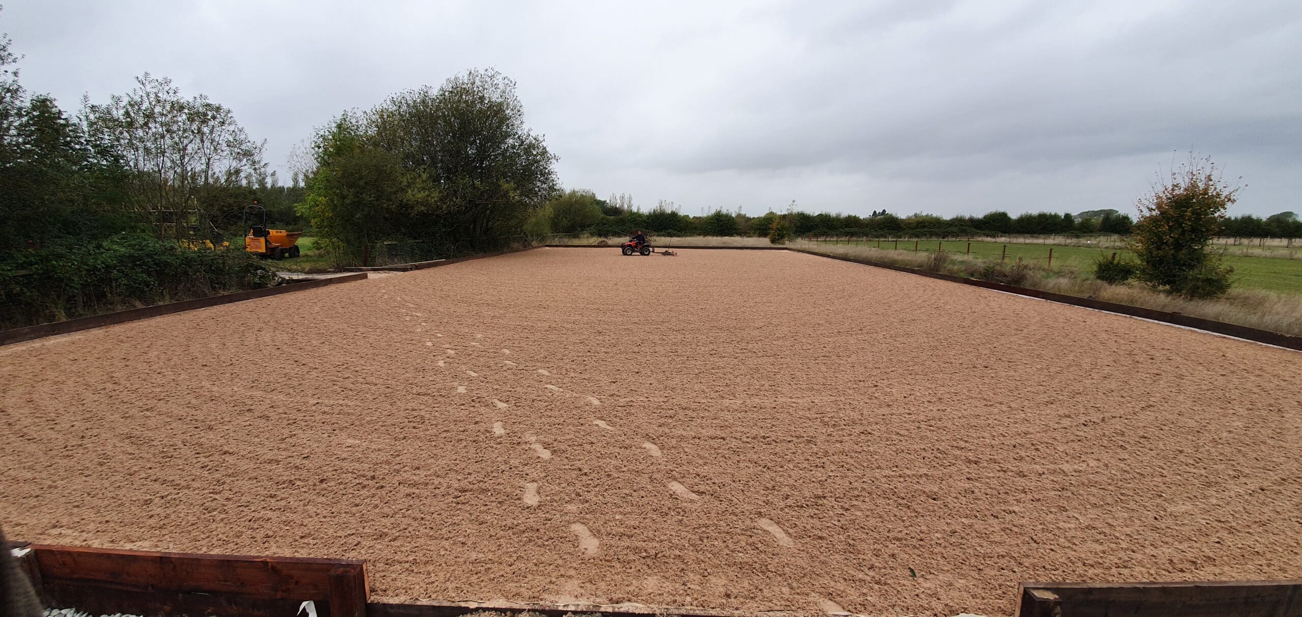 A large, rectangular outdoor equestrian arena with sandy ground and hoofprints. A tractor is seen in the distance preparing the surface. The area is surrounded by lush green trees and open fields under a cloudy sky.