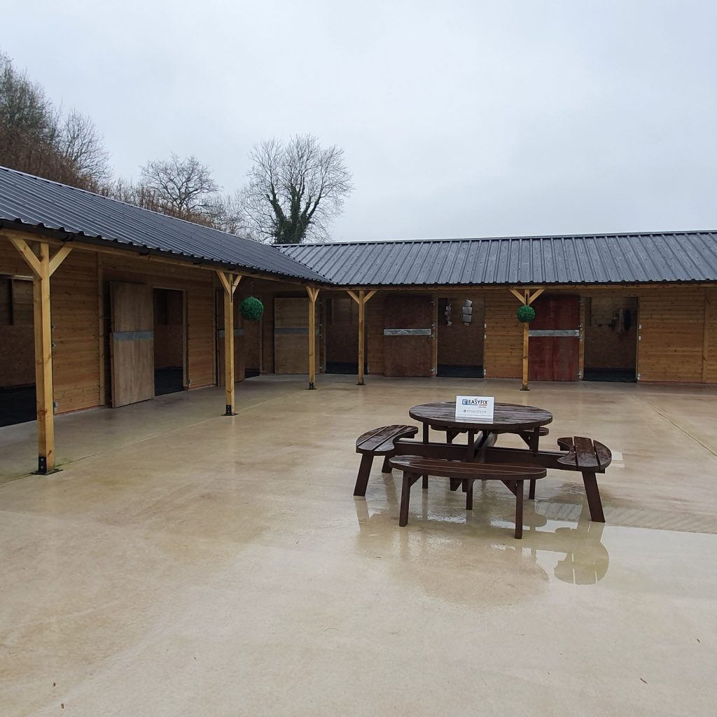 A rainy day scene of a courtyard surrounded by wooden stables. In the center, there is a round picnic table with a white sign on top. The ground is wet, reflecting the overcast sky and surrounding structures.