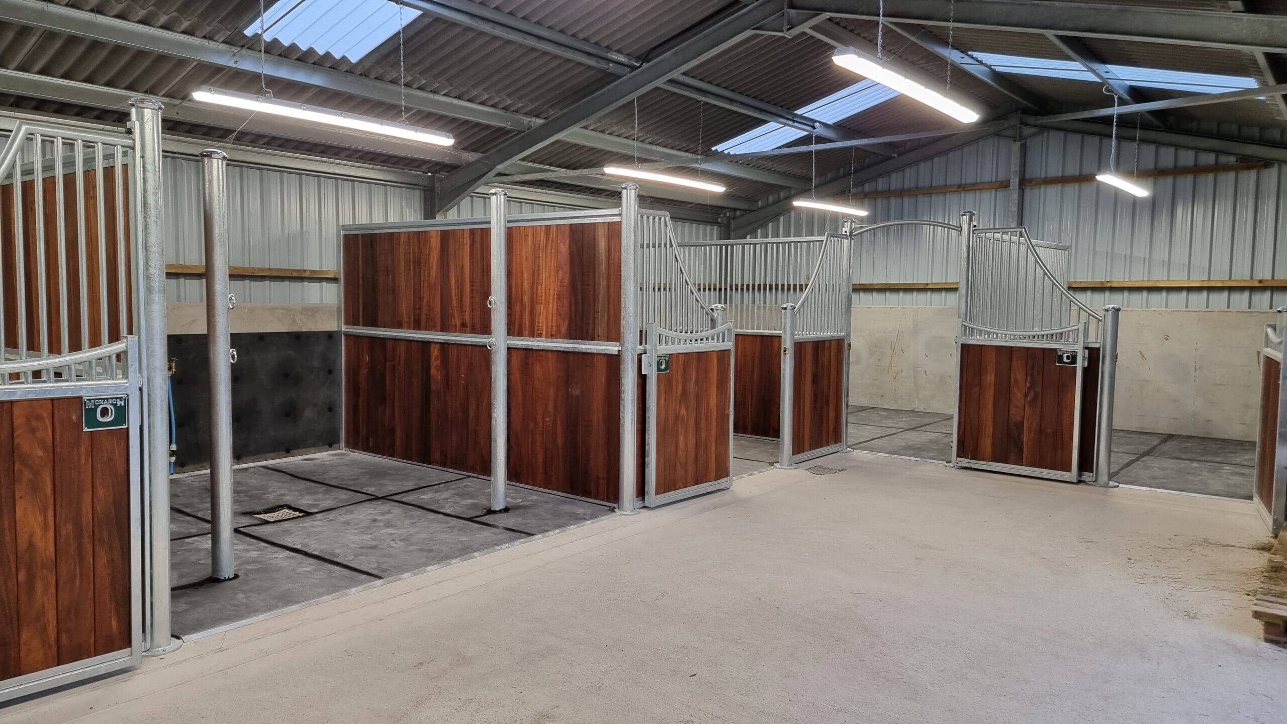 Interior view of a clean, modern stable with open stalls. The walls are made of wood and metal rails, and the floor is concrete. The ceiling features exposed beams and fluorescent lighting.