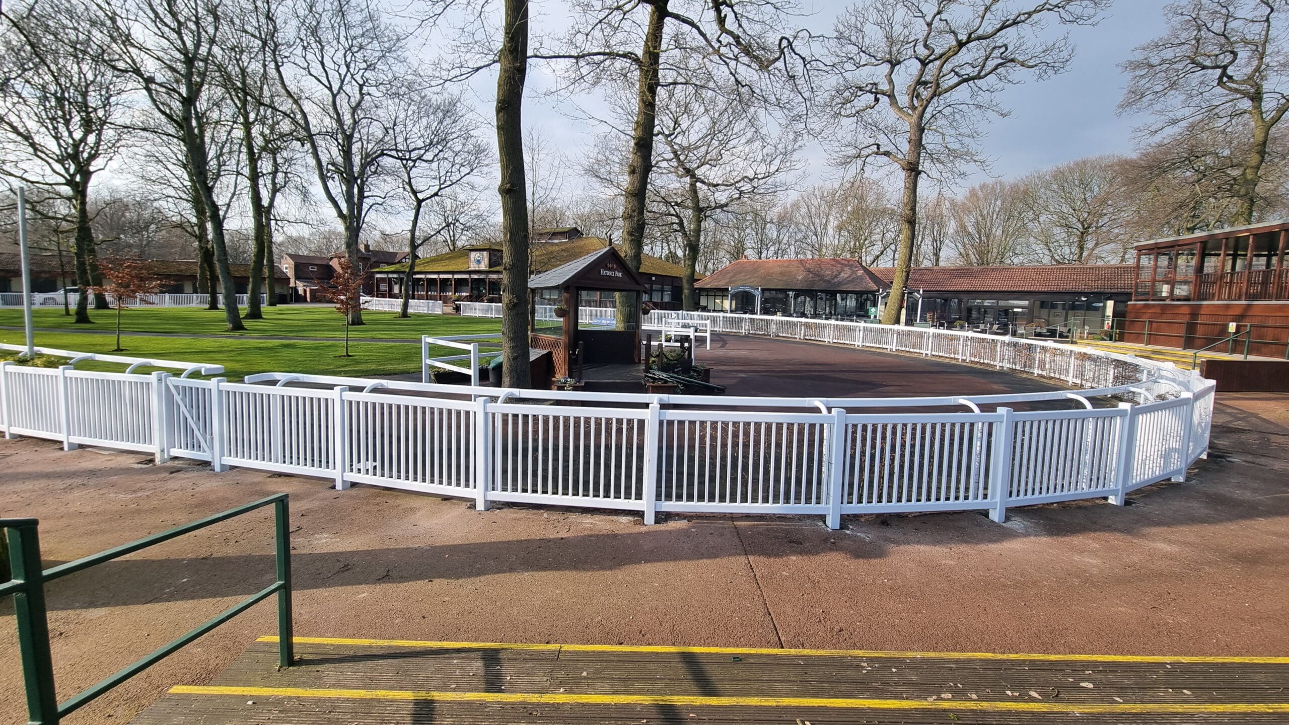 A fenced, circular paddock surrounded by leafless trees. The ground inside is dark and bordered by white rails. In the background, small buildings and green grass are visible under a partly cloudy sky. Steps lead up to the area.