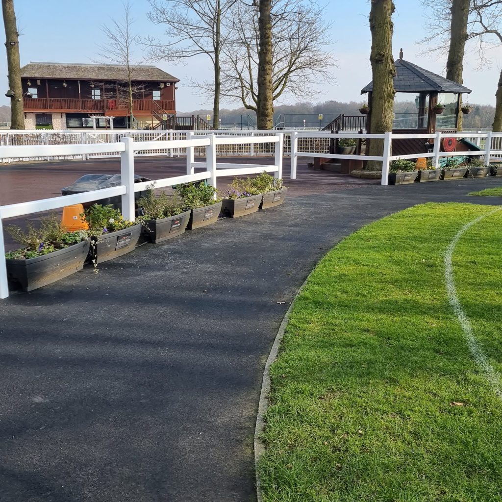 A pathway curves alongside a grassy area with a white fence and planters filled with flowers. Trees line the background near a wooden building, and a small pavilion or gazebo is visible on the right. The scene is bright and peaceful.