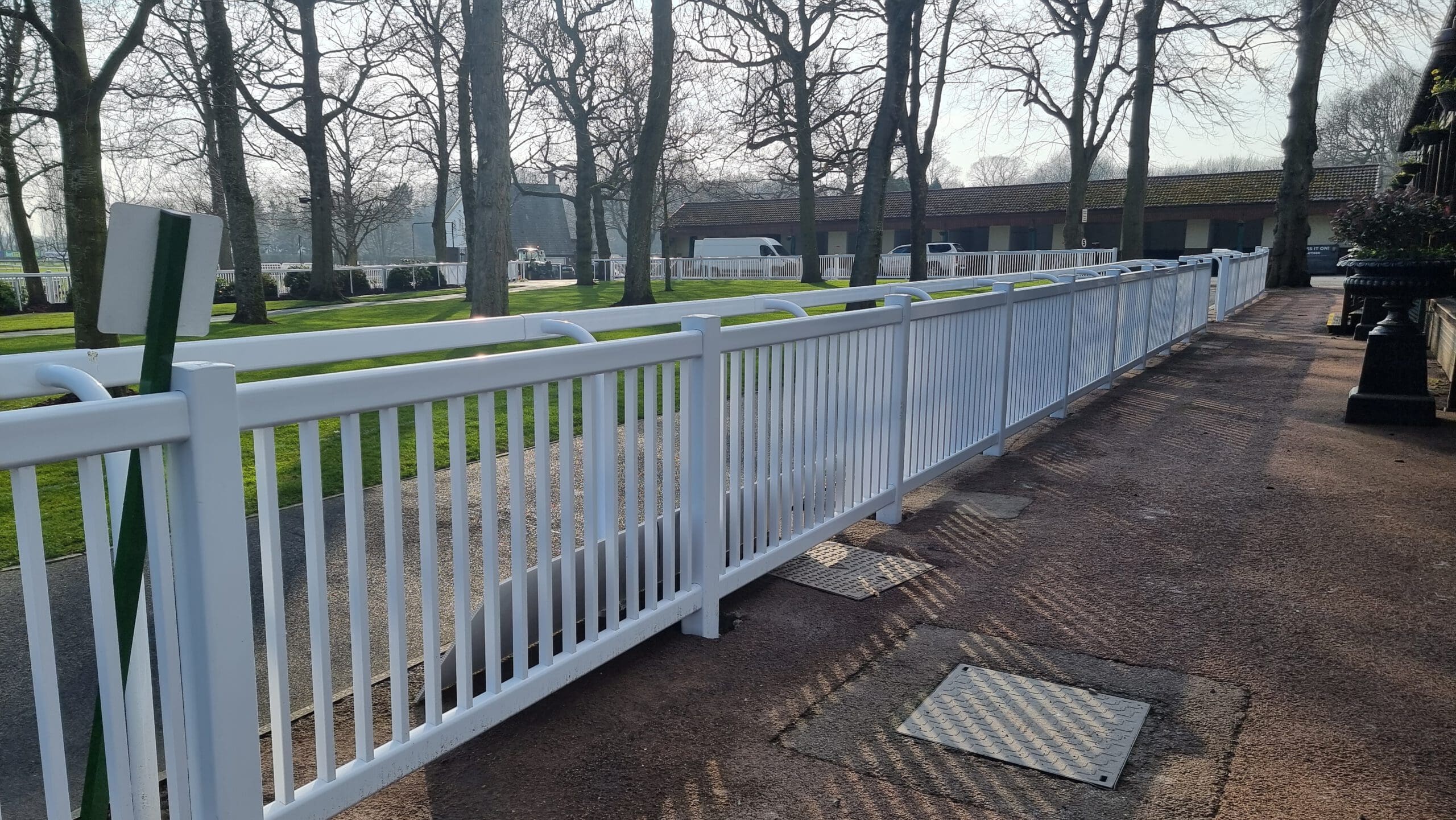 A long white fence lines a paved path, with trees and green grass on the other side. A building with a sloped roof is in the background under a clear sky. Shadows of the trees extend across the ground.