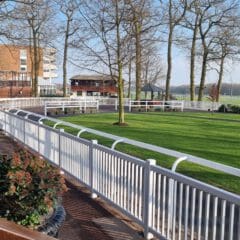 A scenic view of a racetrack with a white railing, lush green grass, and bare trees under a clear blue sky. Buildings and a gazebo are in the background, surrounded by neatly maintained shrubbery along a paved walkway.