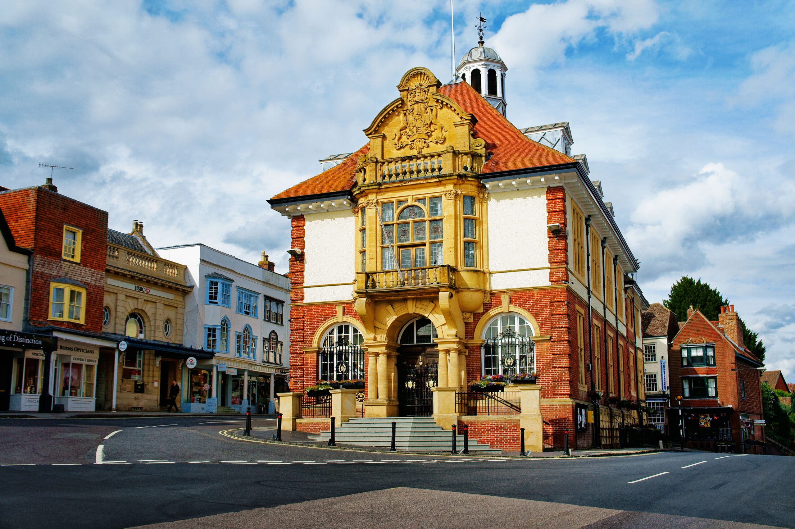 A historic, ornate building with a grand entrance and arched windows stands on a street corner against a cloudy sky. Surrounding it are smaller shops with colorful facades. The scene conveys a blend of architectural styles.